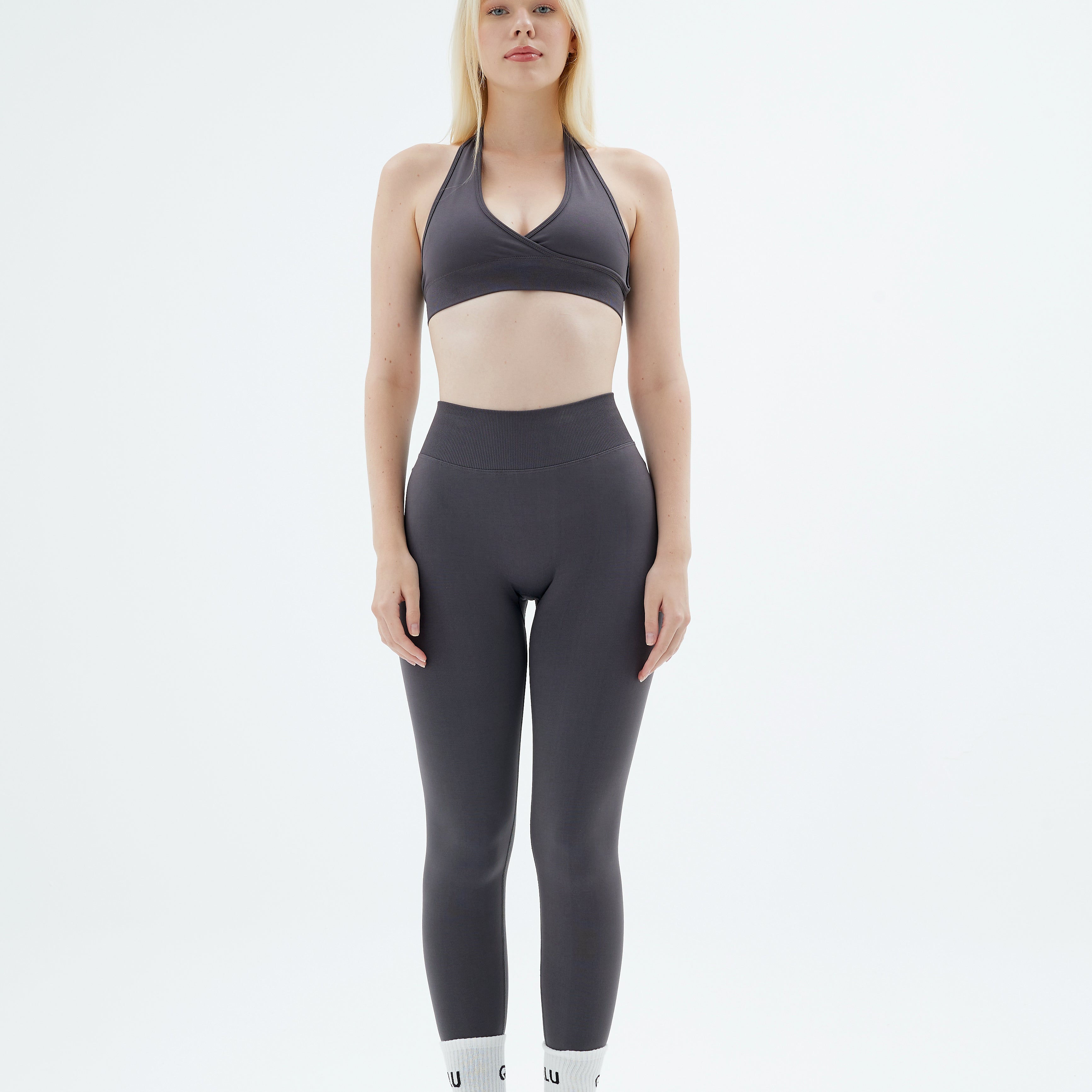 qlu kadın, koyu gri halter yaka spor sütyeni ve tayt, beyaz çorap ve spor ayakkabı giymiş, ayakta duruyor.